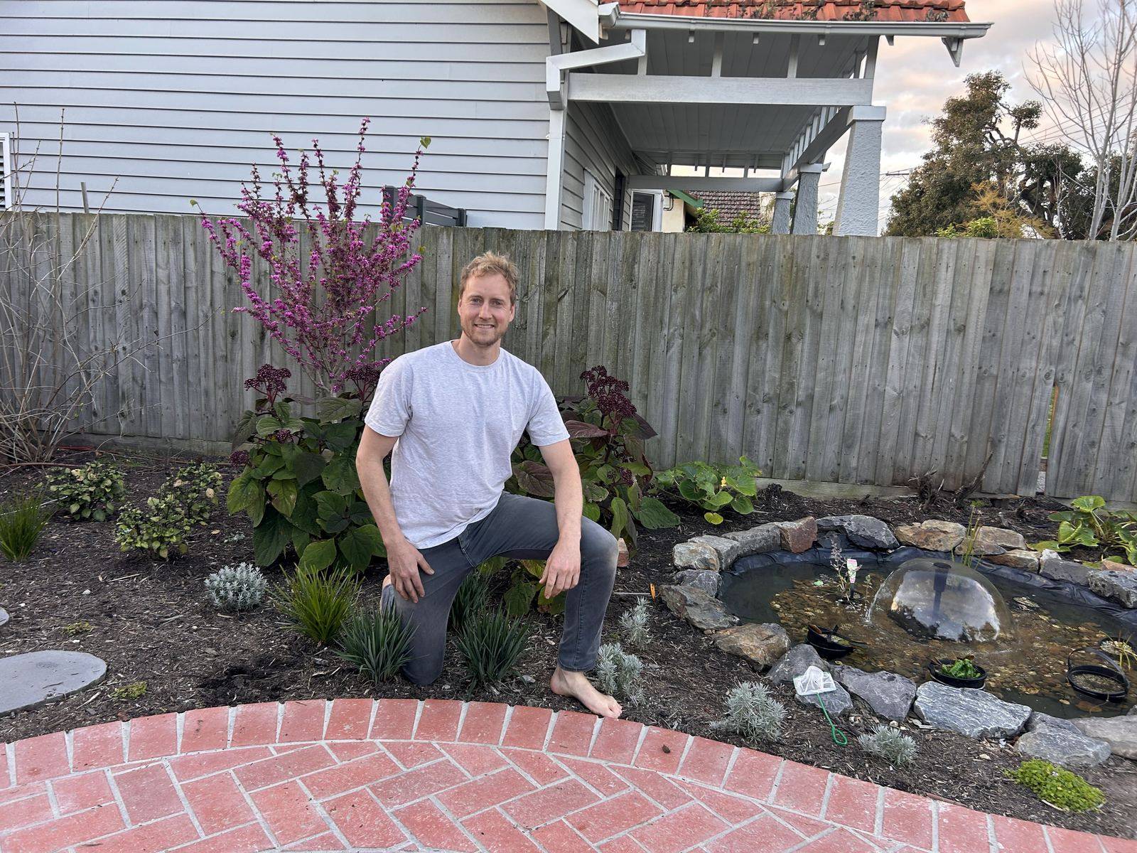 Andy kneeling in a new garden bed beside a rock-edged pond and herringbone brick path